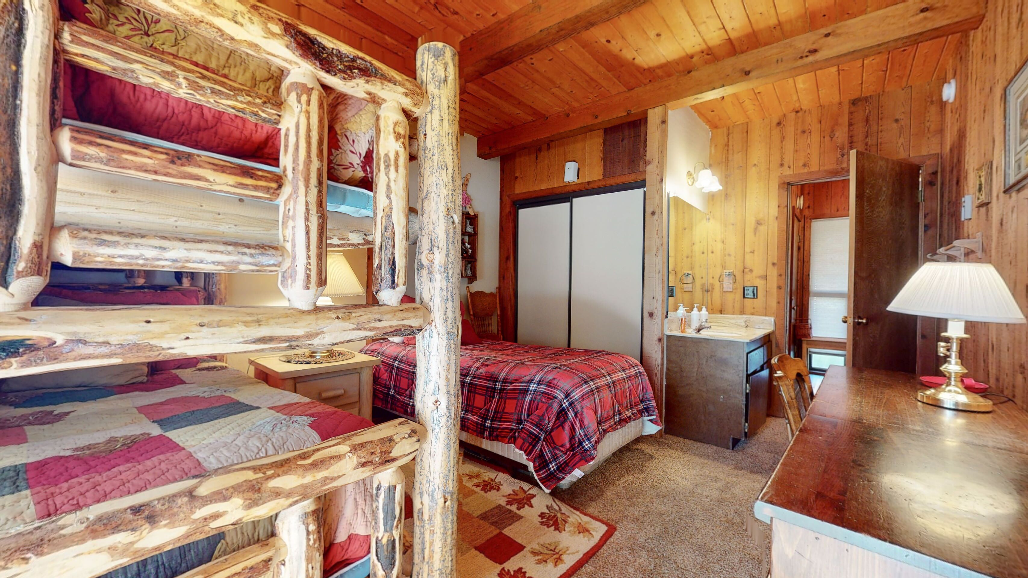 A rustic cabin interior with a wooden bed, a small bedside table, a bathroom with a sink and mirror, and a wooden dresser.