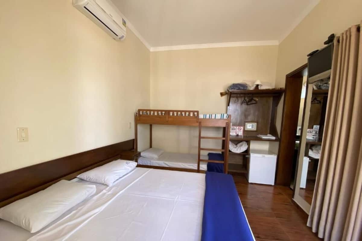 A hotel room with a bunk bed, a single bed, a desk with a chair, a wardrobe, and an air conditioning unit.