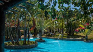 Uma área de piscina com água azul cristalina, cercada por plantas tropicais e uma rede.