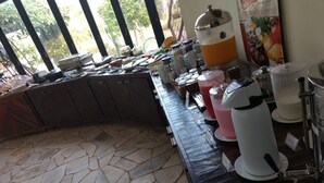 Uma área de buffet de café da manhã com piso de ladrilho, balcão curvo e diversos distribuidores de alimentos e bebidas.