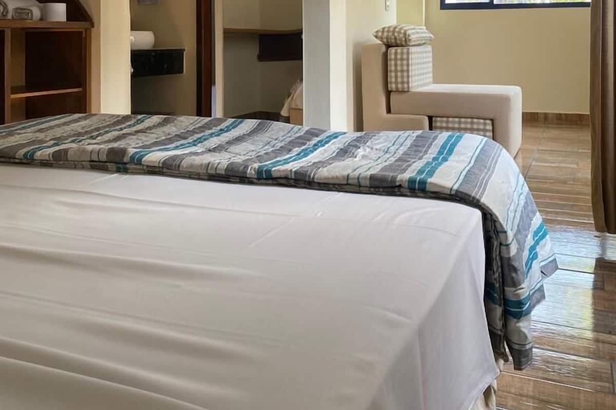 A hotel room with a bed, a TV mounted on the wall, a window with a view of greenery, and a bathroom with a towel hanging on the wall.