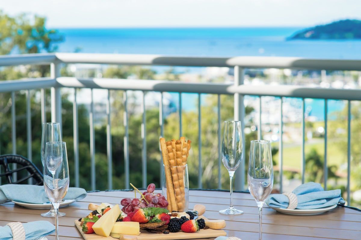 A table with a cheese board, fruits, and crackers, two glasses of water, and a scenic view of the ocean.