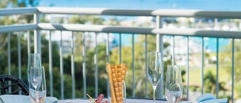 A table with a cheese board, fruits, and crackers, two glasses of water, and a scenic view of the ocean.