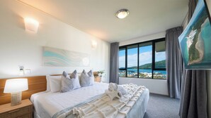 A hotel room with a large bed, a nightstand, a lamp, a painting, and a view of the sea. - Paradise Palms (Airlie Beach)