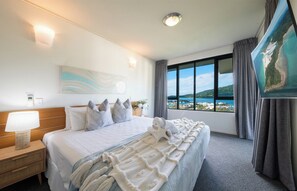 A hotel room with a large bed, a nightstand, a lamp, a painting, and a view of the sea. - Paradise Palms (Airlie Beach)