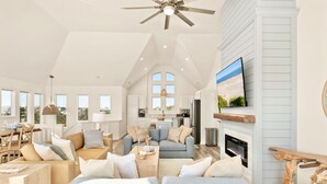 A spacious living room with a high ceiling, a fireplace, and a television mounted on the wall. - Oceanfront Luxury Panoramic 6BR, Pool, Hot Tub, Game Room, Surf House OBX (Salvo)