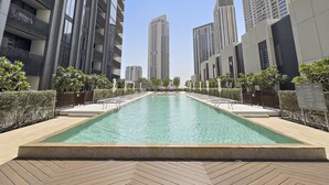 A modern urban pool area with a long rectangular pool, surrounded by high-rise buildings and well-maintained greenery.