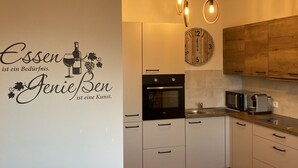 Una cocina moderna con armarios blancos, horno empotrado, microondas y encimera de madera. Se observa un reloj de pared y una lámpara en el techo.