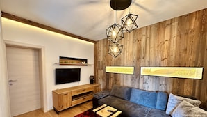 Una sala moderna con pared de madera, televisor de pantalla plana, mueble bajo para televisión de madera, sofá gris, alfombra roja y lámparas colgantes geométricas.