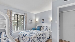 Une chambre avec un lit superposé, un lit simple, une fenêtre avec des stores, une décoration en forme de bouée de sauvetage et un tableau encadré au mur.