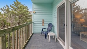 Un balcon avec un mur extérieur bleu, une balustrade en bois, une chaise bleue, un petit tabouret blanc et une porte-fenêtre coulissante donnant sur un espace extérieur.