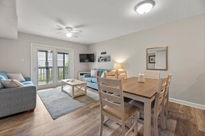 A modern living room with a dining area, a ceiling fan, and a TV mounted on the wall. - The Hawk's Nest: Renovated Condo | 700ft. to Beach (Corolla)