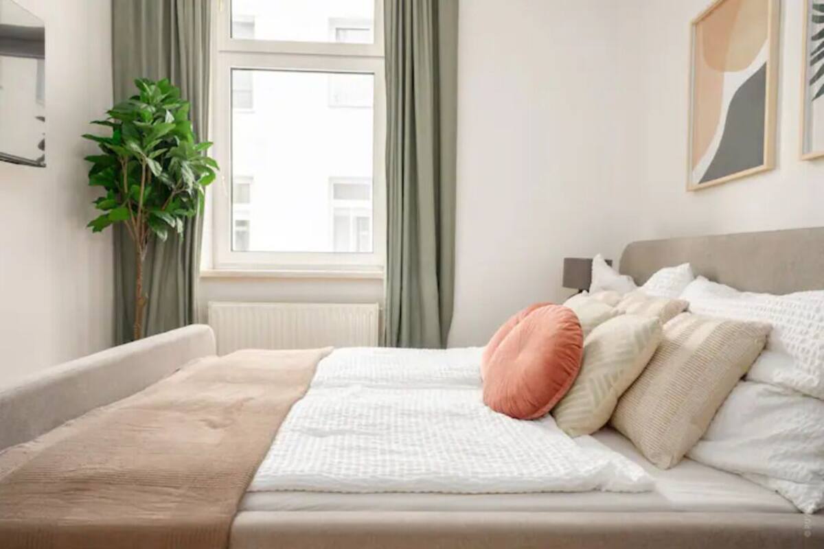 A neatly made bed with a beige blanket and pillows, a green plant beside a window, and a framed picture on the wall.