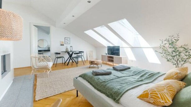 A modern loft bedroom with a skylight, a bed with green and yellow pillows, a fireplace, and a dining area with a round table and chairs. - Designer Apartments - Near Reumannplatz (Vienna)