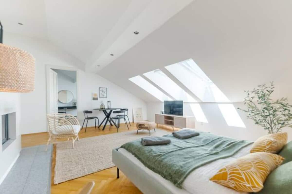 A modern loft bedroom with a skylight, a bed with green and yellow pillows, a fireplace, and a dining area with a round table and chairs.