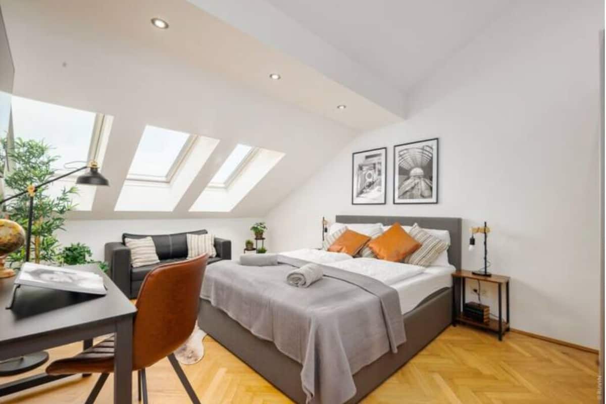 A modern bedroom with a skylight, a bed with pillows, a desk with a chair, and a small table with a lamp.