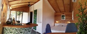 Ein Holzblockhaus-Interieur mit Küchenbereich, einer Holzauslage, einem Korb und Weinflaschen. Ein Bett, ein blauer Stuhl und ein kleines gerahmtes Bild sind an der Wand zu sehen.