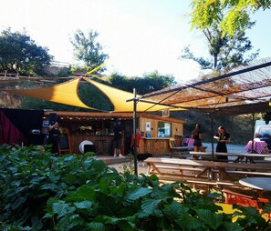 Un café al aire libre con mesas y bancos de madera, un toldo amarillo y un edificio rústico de madera con una zona de bar.