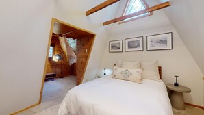 Une chambre avec un plafond en pente, un lit avec une literie blanche, une table de chevet et trois tableaux encadrés accrochés au mur.