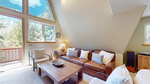 Un salon chaleureux avec un canapé en cuir marron, une table basse en bois et un fauteuil blanc. Il y a un balcon avec une balustrade en bois et une vue sur les arbres.