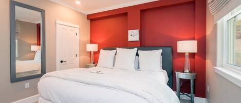 A bedroom with a large bed, two bedside tables with lamps, a mirror, and a red accent wall.