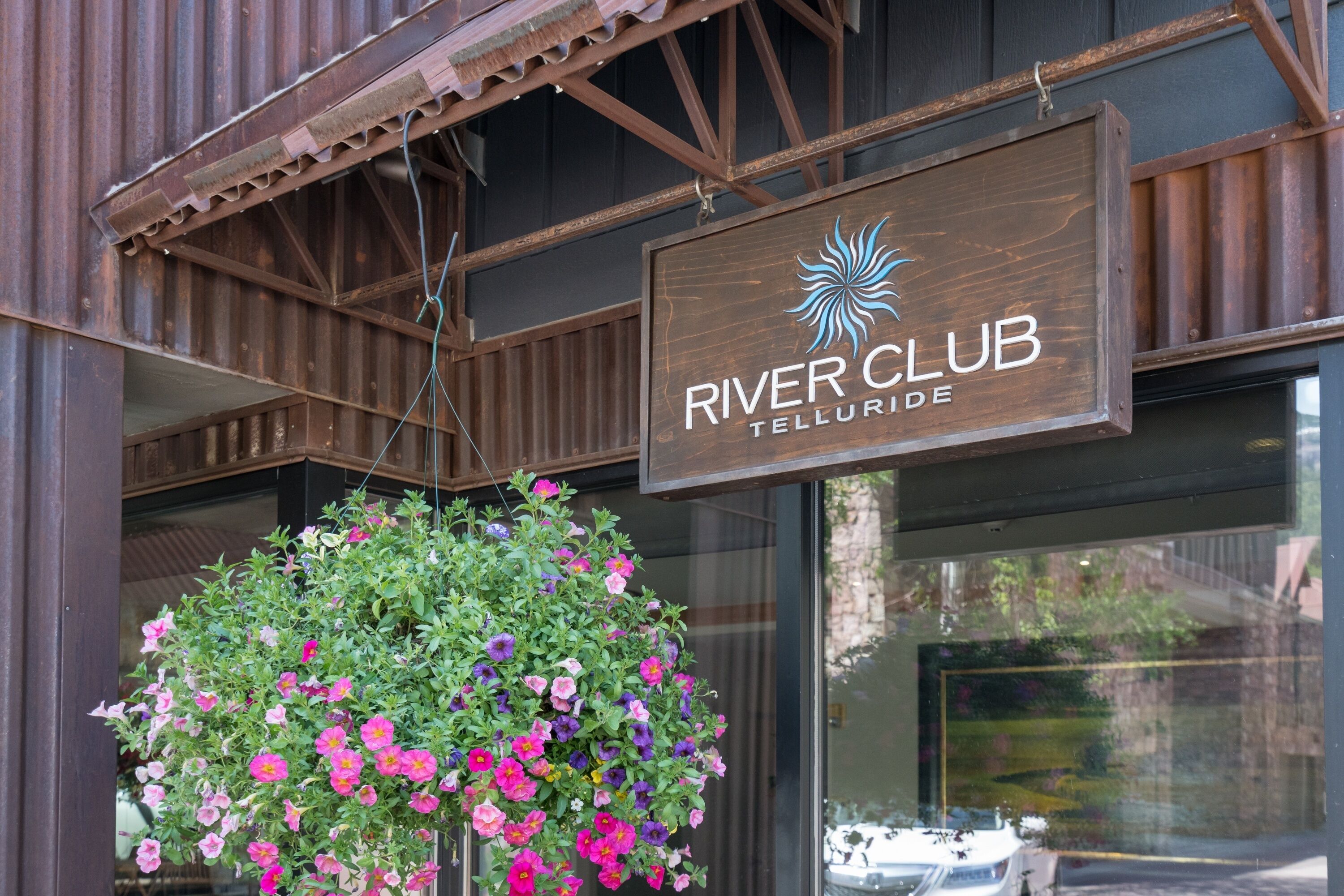 Un letrero de madera con el texto 'RIVER CLUB' y 'TELLURIDE', una maceta colgante con flores rosas y púrpuras, y un toldo metálico encima.