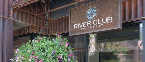 Un letrero de madera con el texto 'RIVER CLUB' y 'TELLURIDE', una maceta colgante con flores rosas y púrpuras, y un toldo metálico encima.