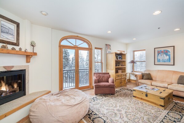 Cozy up by the fireplace in this inviting living room with a balcony offering views of snow-covered trees.