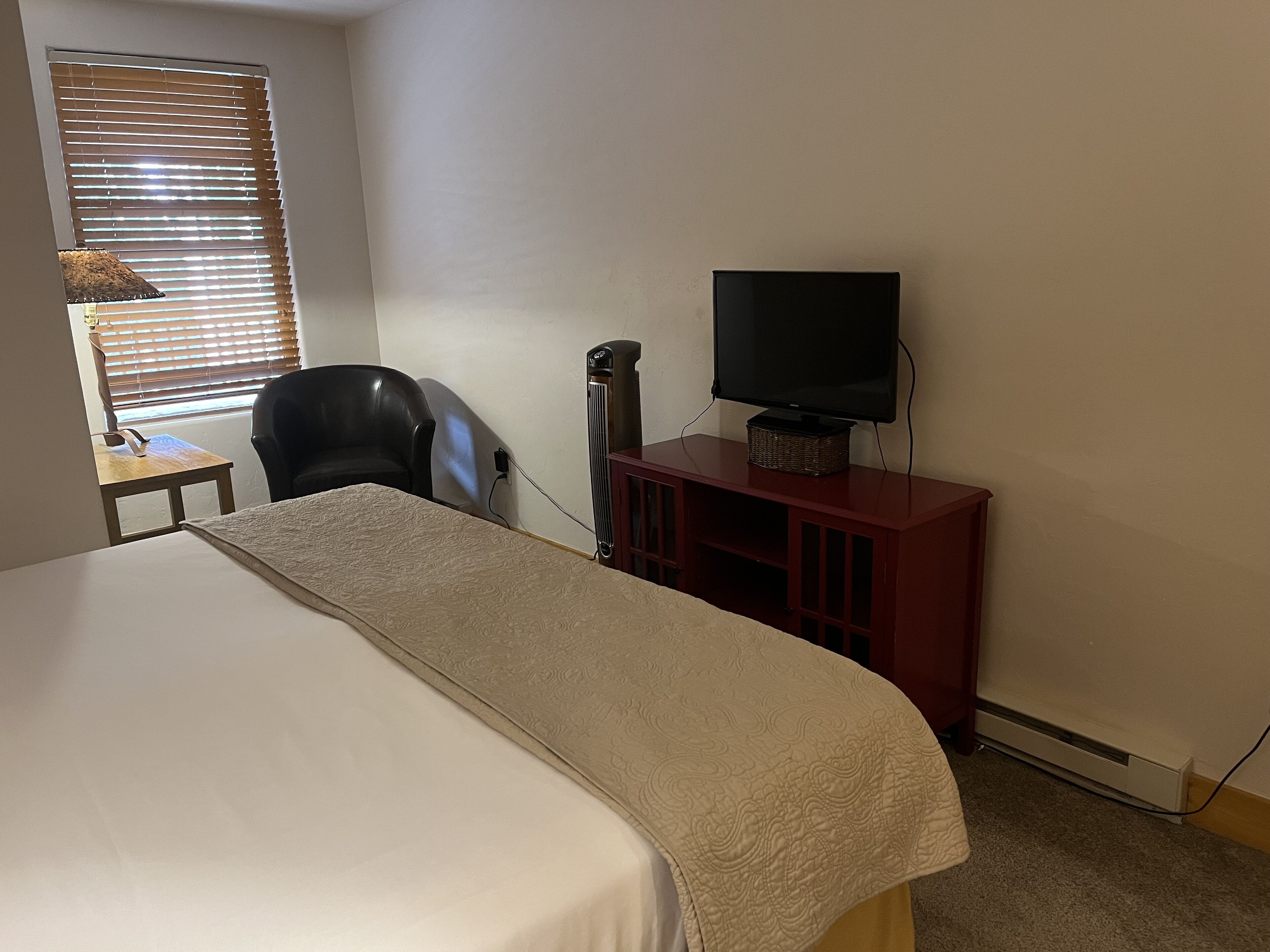 A hotel room with a bed, a television on a wooden stand, a chair, and a window with blinds.