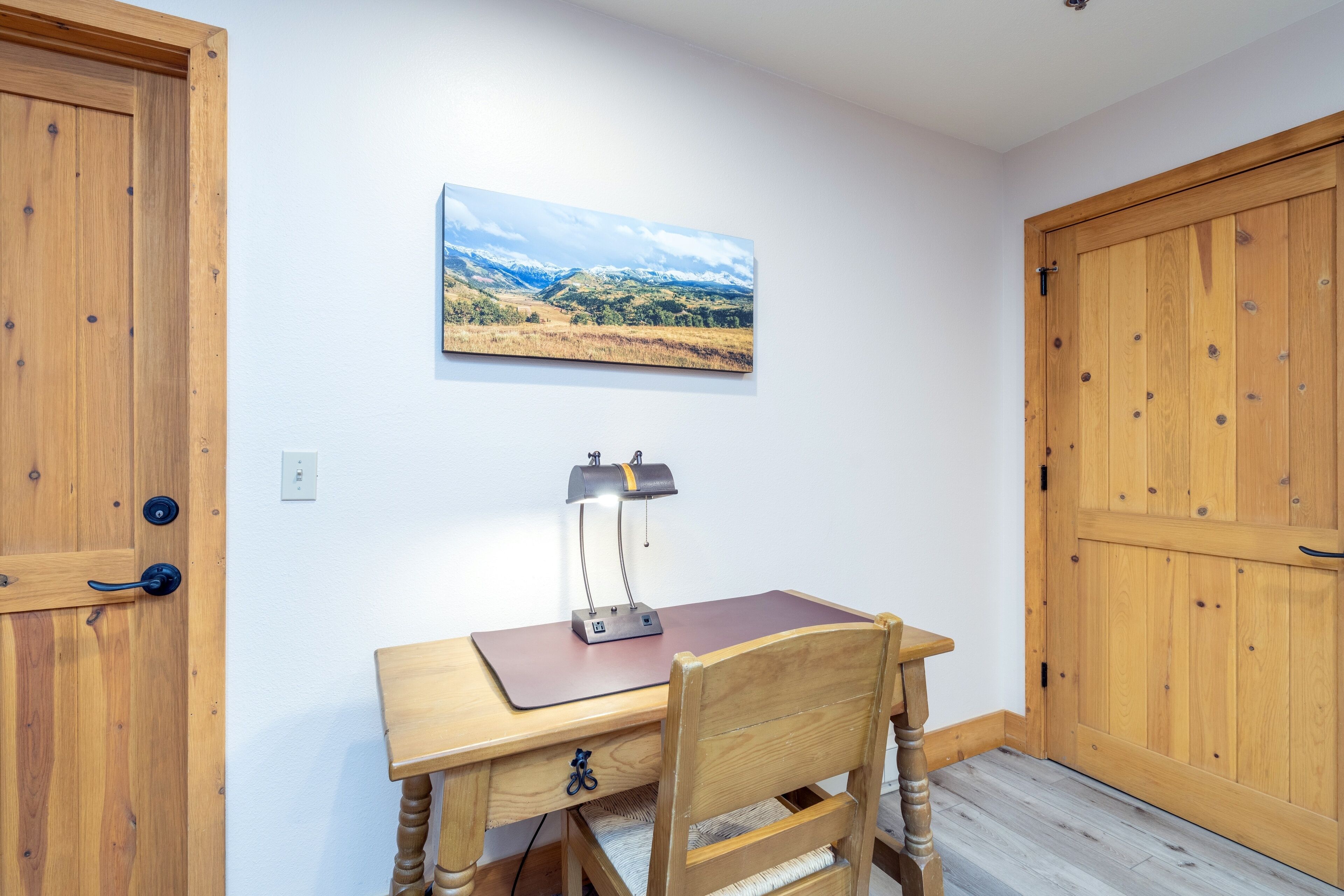 Un escritorio de madera con una lámpara, una silla y un cuadro de paisaje en la pared.
