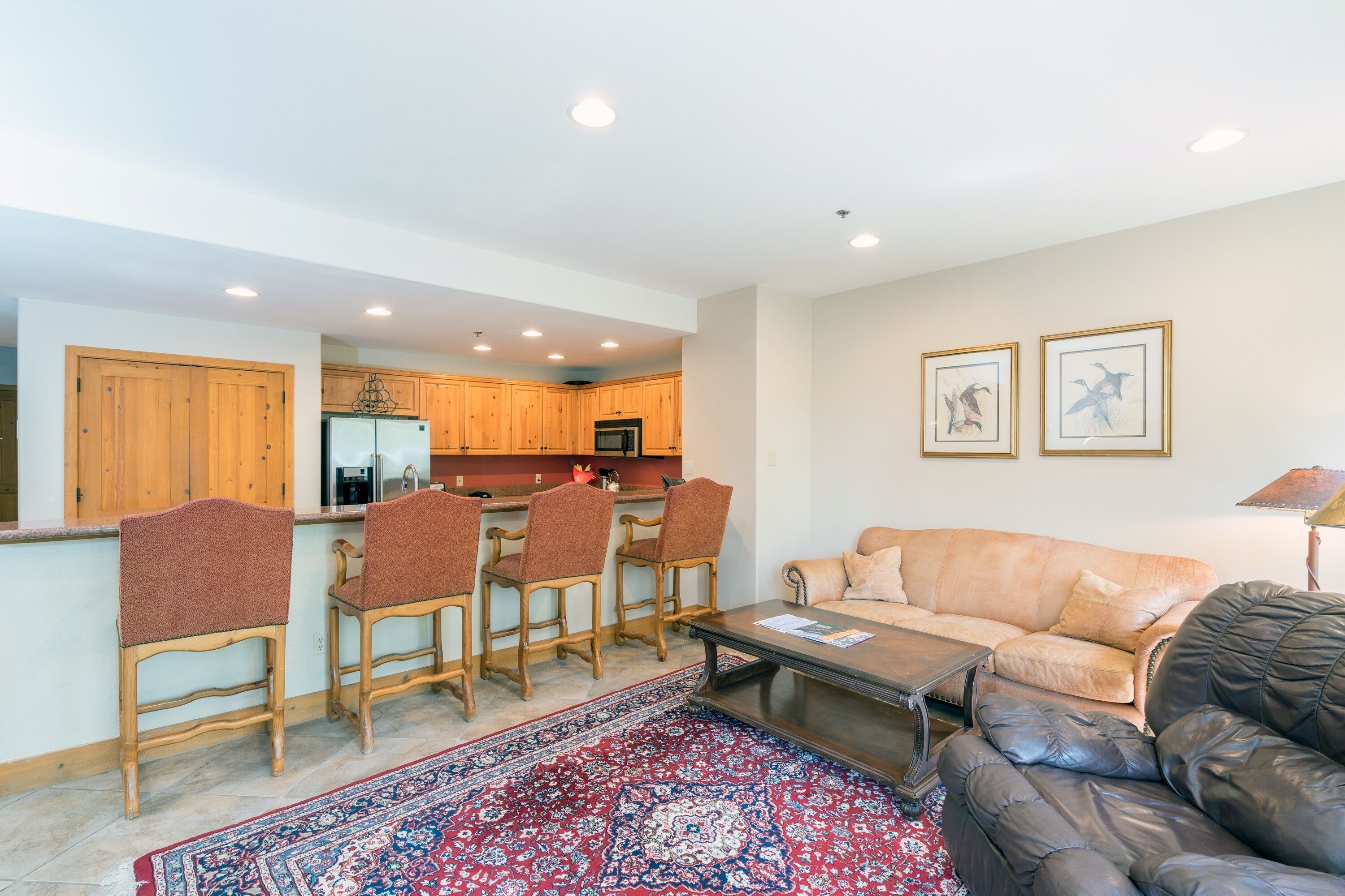 A spacious living area with a sofa, a coffee table, and a patterned rug. A kitchen with wooden cabinets and a refrigerator is visible in the background.