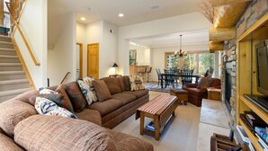 A spacious living room with a large sofa, a coffee table, and a fireplace. There is a staircase leading to another level, and a dining area with a table and chairs. - 1952 Northstar 3 Bedroom Townhouse by RedAwning (Keystone)