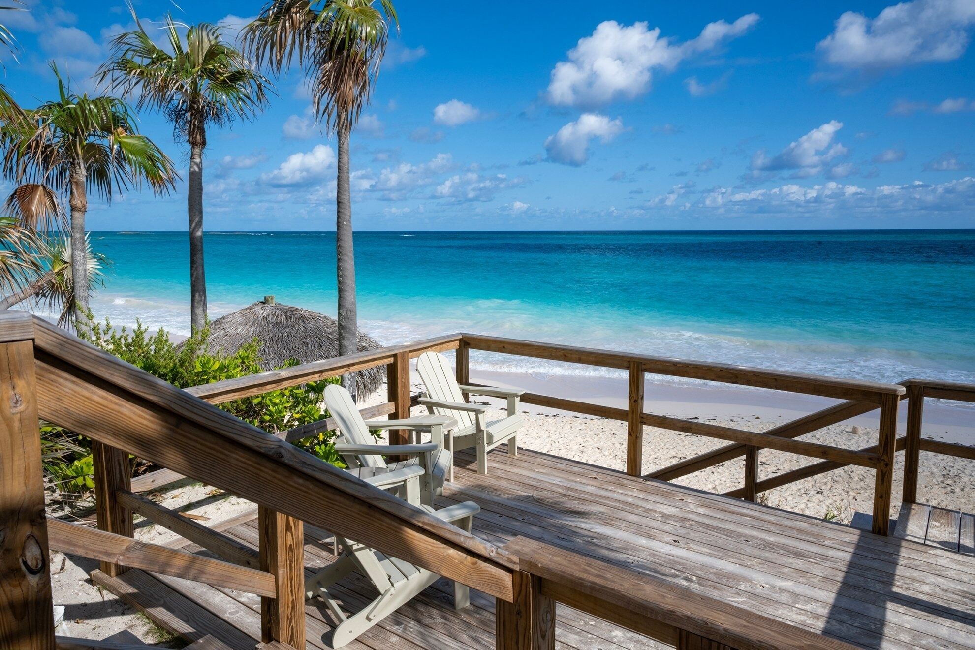 Une terrasse en bois, dotée de chaises de plage et surmontée d’une structure à toit de chaume, offre une vue sur une plage aux eaux turquoise et limpides, bordée de palmiers.