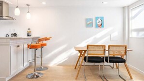 A modern kitchen with white cabinets, a stainless steel range hood, and a bar stool with orange seats. There is a wooden dining table with two wicker chairs. Two framed pictures are hung on the wall.