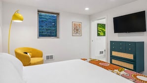 A modern hotel room with a bed, a yellow armchair, a large window with a view, a flat-screen TV, and a decorative cabinet.