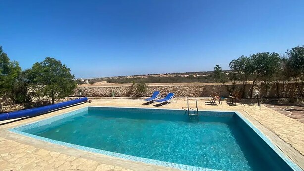 Ein rechteckiger Swimmingpool mit klarem, blauem Wasser, umgeben von einer Steindeck. Zwei blaue Liegestühle und ein kleiner Tisch mit einem Stuhl stehen bereit. Im Hintergrund sind Bäume und eine Steinmauer zu sehen.