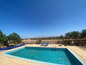 Une piscine rectangulaire à l’eau d’un bleu limpide, entourée d’une terrasse en pierre. On y trouve deux chaises longues bleues et une petite table avec une chaise. La piscine est située devant un décor composé d’arbres et d’un mur en pierre.
