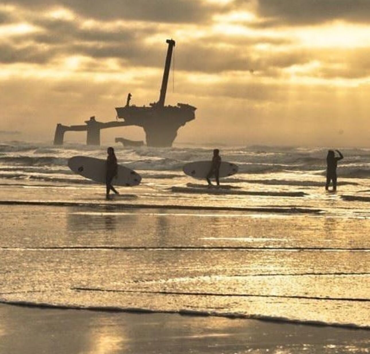 A beach scene with surfers and a large shipwreck in the background.