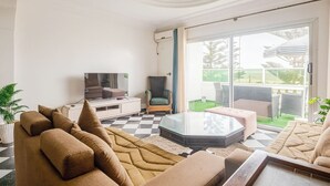 Ein Wohnzimmer mit einer Couch, einem Glastisch, einem Fernseher und einem Sessel. Ein großes Fenster bietet Blick auf einen Garten.
