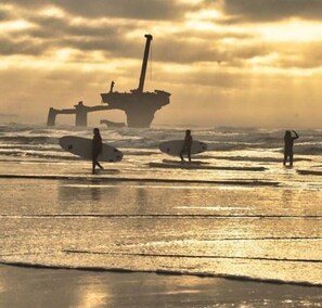 Une scène de plage avec des surfeurs et, au loin, les restes d’un grand navire échoué.
