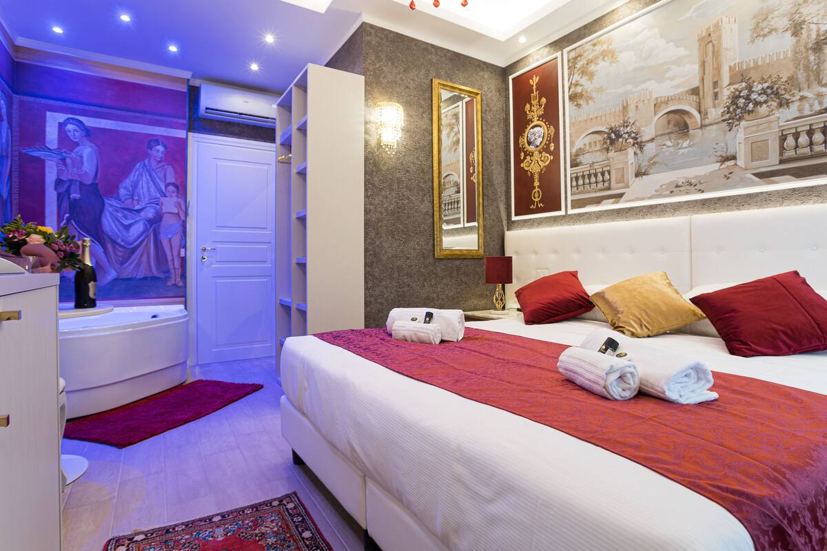 Habitación de hotel con una cama grande, almohadas rojas y doradas, un adorno de pared rojo y dorado, un armario blanco y un baño con un tapete púrpura.