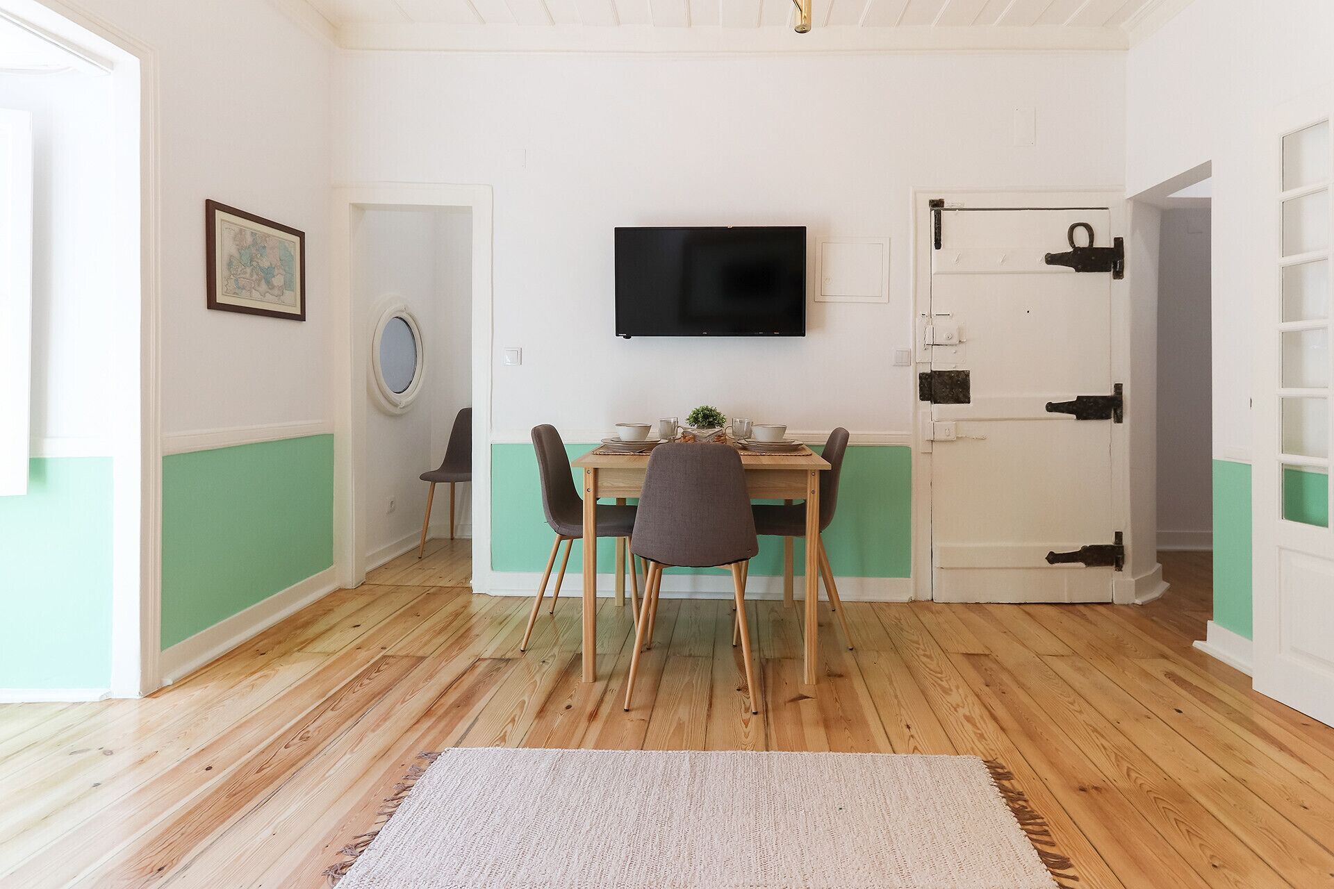 A dining area with a wooden table, four chairs, a television above the table, and a door with a handle.