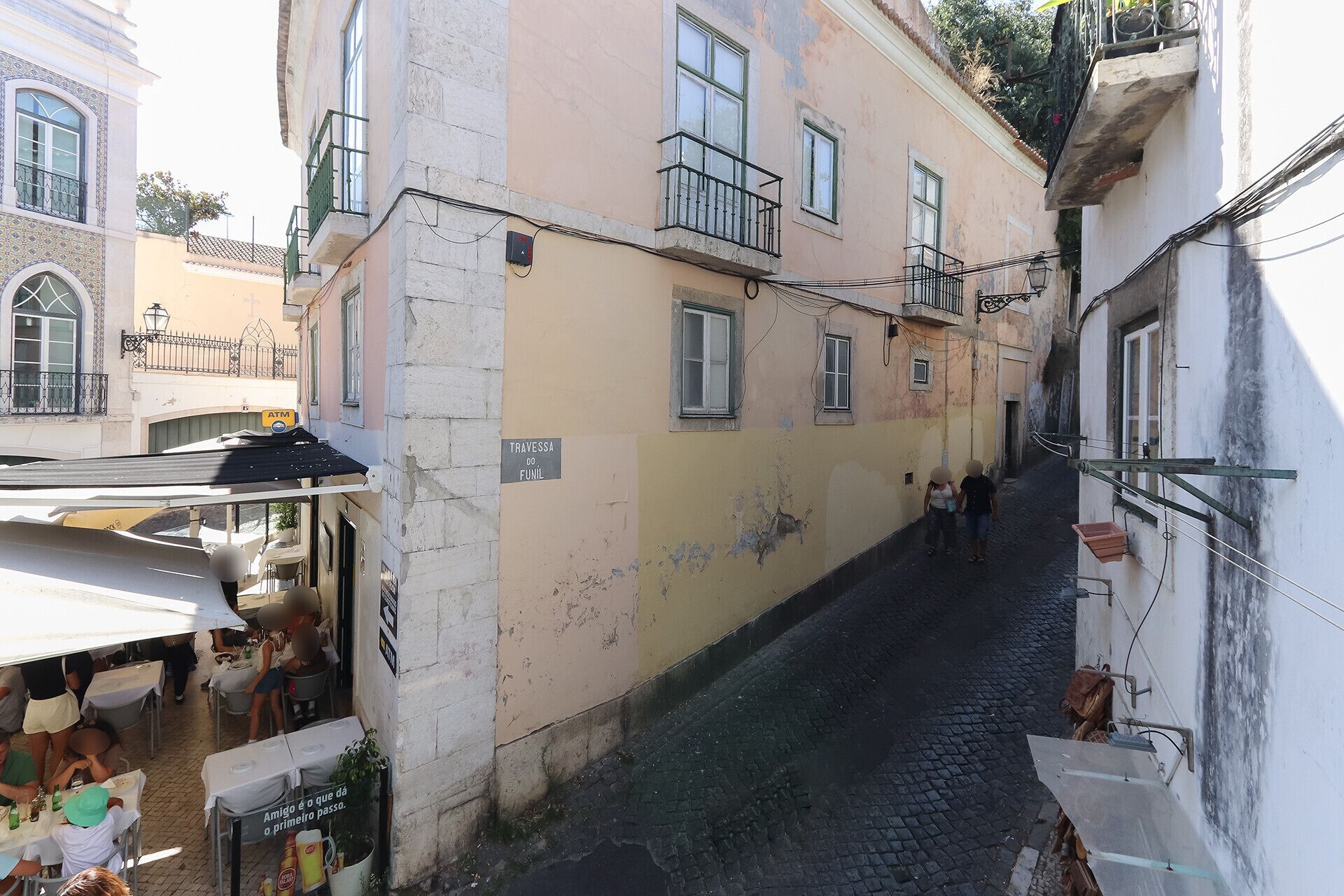 A narrow street with buildings on either side, a visible ATM, and outdoor seating for a café.