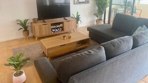 A modern living room with a flat-screen TV on a wooden stand, a grey sofa, and two potted plants.