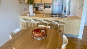 A modern kitchen with a central island, white bar stools, and a dining table with a fruit basket.