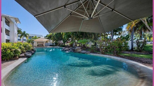 A clear swimming pool with a shaded area, surrounded by tropical plants and a building in the background.