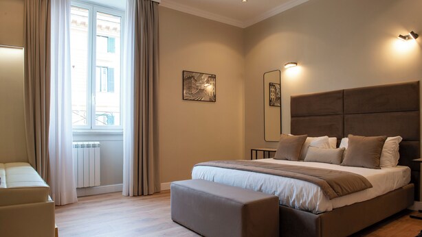 A modern bedroom with a large bed, a bench, a chair, and a view of the street through a window. - HOTEL AD Suites Nazionale (Rome)