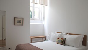 Un dormitorio con cama, mesita de noche, una silla, ventana y un cuadro enmarcado en la pared.
