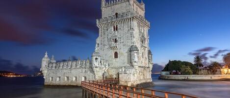 A historic stone castle with a prominent tower, a wooden walkway leading to it, and a calm body of water in the foreground.