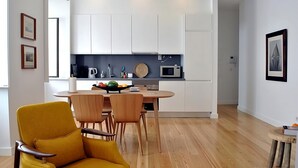 Une cuisine moderne avec des meubles blancs, un four à micro-ondes et un panier de fruits sur le comptoir. On aperçoit une table à manger avec des chaises au premier plan.